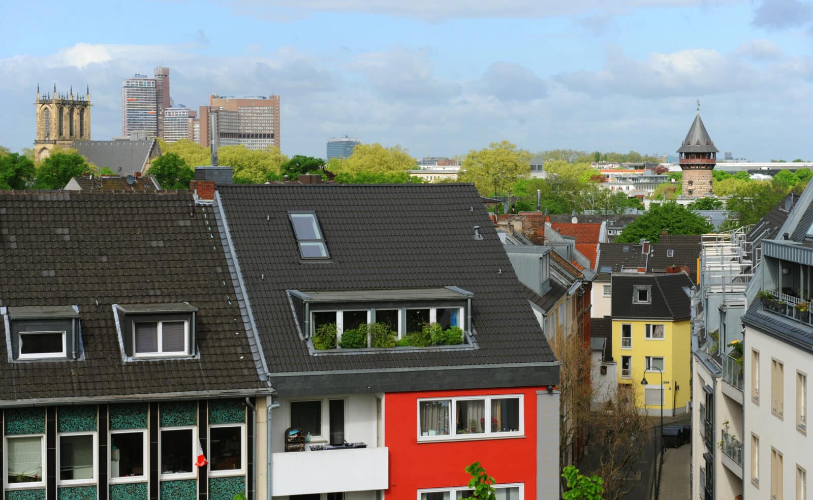 Ein Stadtbild mit farbenfrohen Wohnhäusern im Vordergrund – was vielleicht die Frage aufwirft: „Soll ich vor dem Verkauf mein Haus in Köln renovieren?“ –, grünen Bäumen und historischen Kirchtürmen im Hintergrund unter einem teilweise bewölkten Himmel.