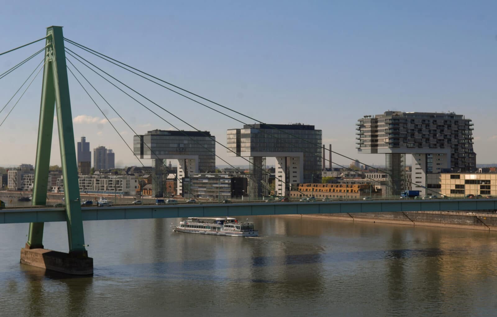 Eine Flussszene mit einer modernen grünen Hängebrücke im Vordergrund, einem weißen Passagierboot auf dem Wasser und drei markanten kranförmigen Gebäuden entlang des Flussufers im Hintergrund unter einem klaren blauen Himmel.