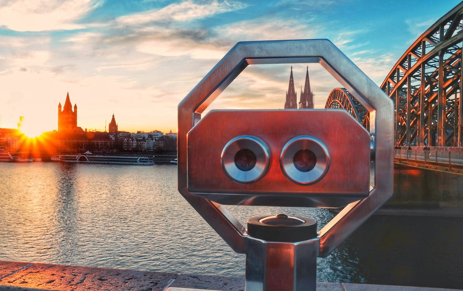 Ein münzbetriebener Binokularbetrachter auf einer Brücke überblickt einen Fluss bei Sonnenuntergang, mit der Skyline einer Stadt und den Türmen einer Kathedrale im Hintergrund und einer gewölbten Eisenbrücke auf der rechten Seite.