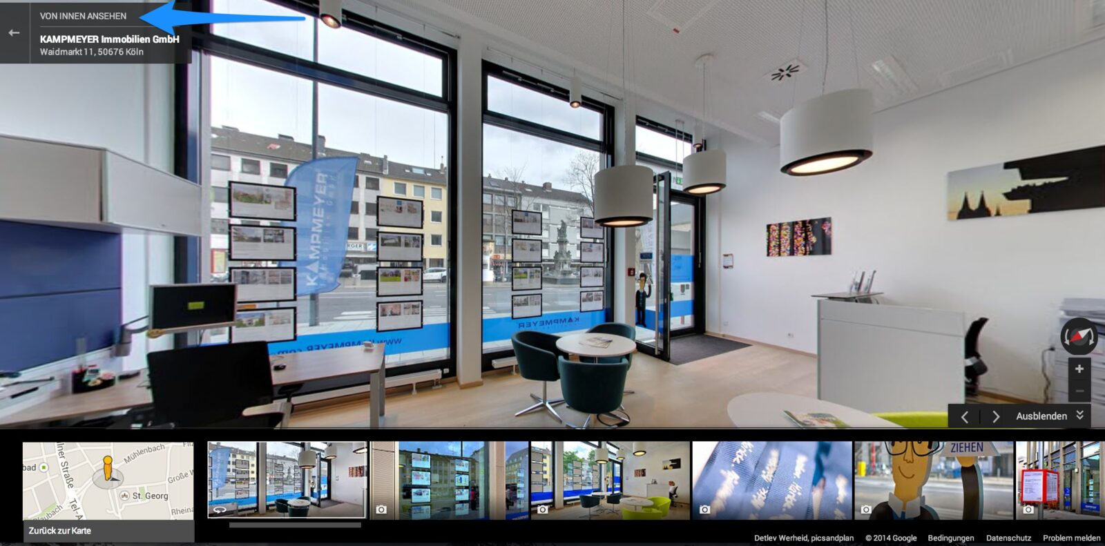 Ein helles Immobilienbüro mit großen Fenstern, modernen Möbeln und ausgestellten Immobilienangeboten. Computermonitore stehen auf weißen Schreibtischen, und von der Decke hängen Pendelleuchten. Eine Tür führt nach draußen auf die Straße.