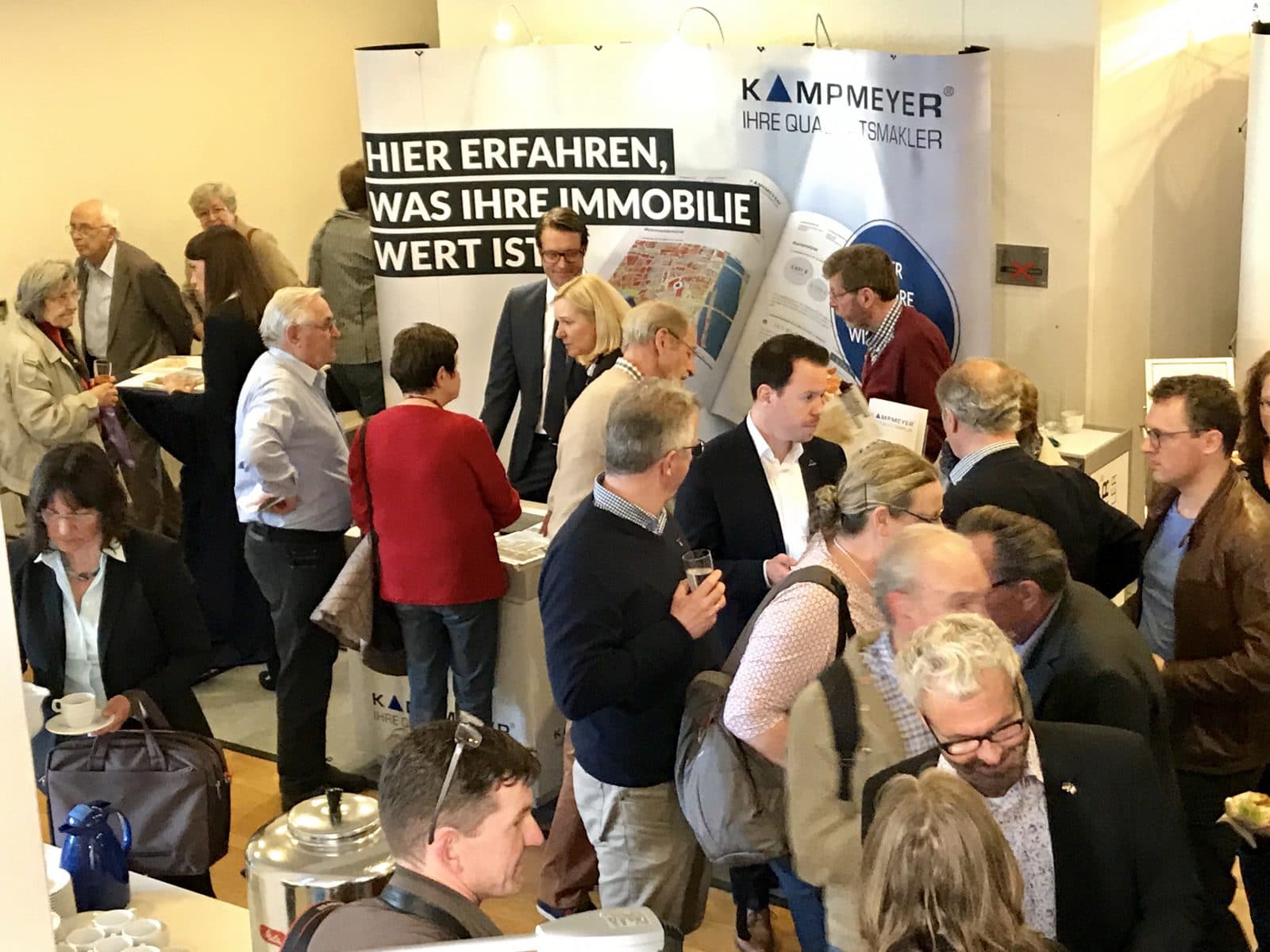 Eine Gruppe von Menschen knüpft Kontakte und unterhält sich auf einer Immobilienveranstaltung, die in der Nähe von Ausstellungsständen mit deutschsprachigen Bannern stattfindet. Die Veranstaltung findet in einem Innenraum statt, in dem Informationsmaterial und Kaffee auf den Tischen stehen.
