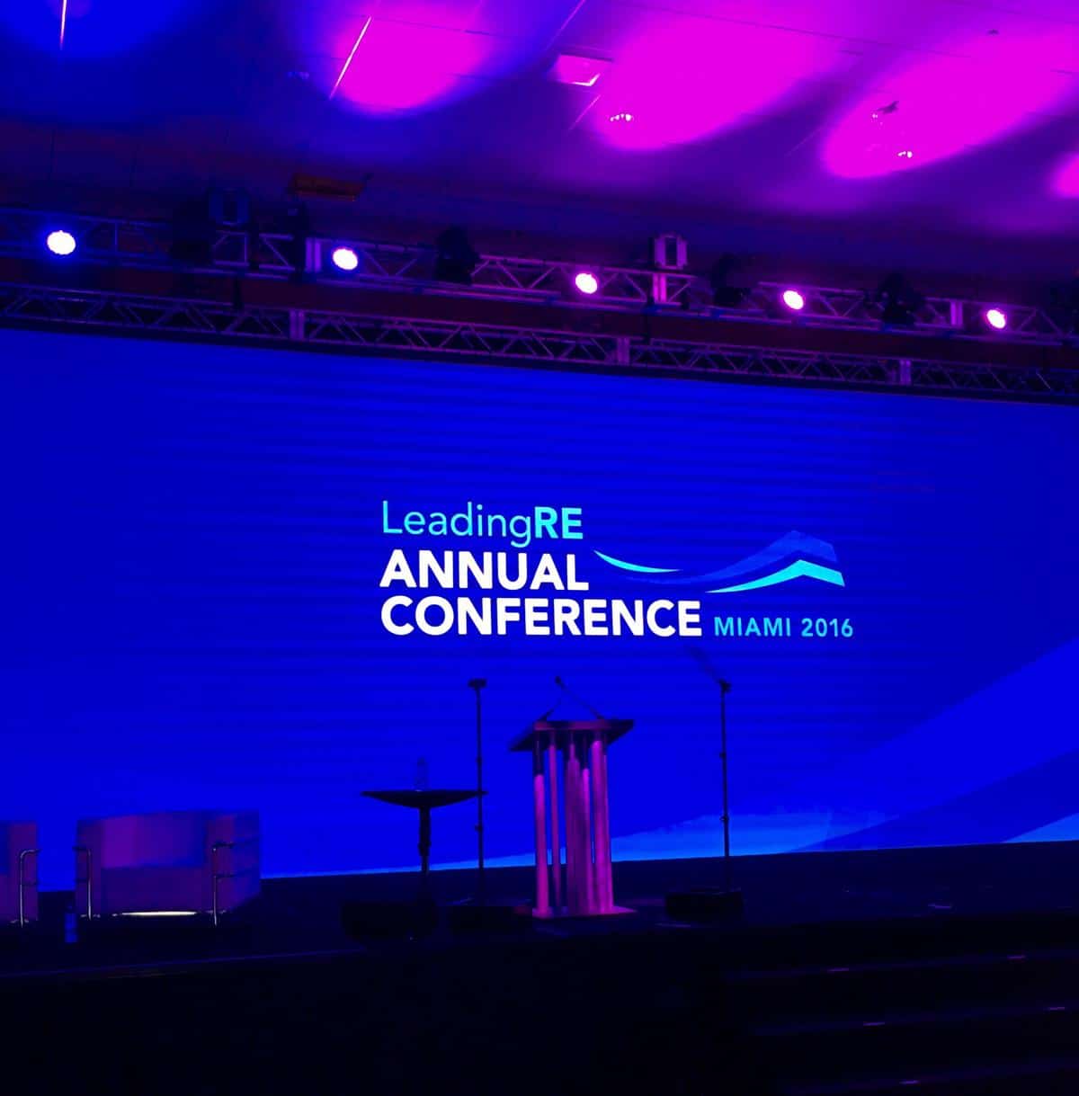 Auf einer mit lila und blauen Lichtern beleuchteten Bühne ist ein großer Bildschirm mit der Aufschrift LeadingRE Annual Conference Miami 2016 zu sehen. Vor dem Bildschirm sind ein Podium und Mikrofone aufgebaut.