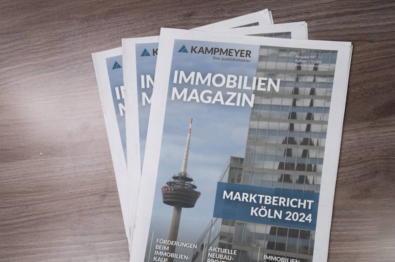Drei Exemplare einer Immobilienzeitschrift mit dem Titel IMMOBILIEN MAGAZIN mit dem Foto eines Fernmeldeturms und eines modernen Glasgebäudes auf dem Umschlag, ausgelegt auf einer Holzfläche.