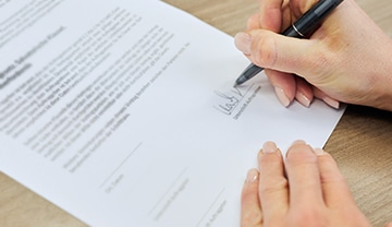 Eine Person hält einen Stift in der Hand und unterschreibt ein gedrucktes Dokument auf einem Holztisch. Nur die Hände und ein Teil des Papiers mit dem getippten Text und den Unterschriftslinien sind sichtbar.