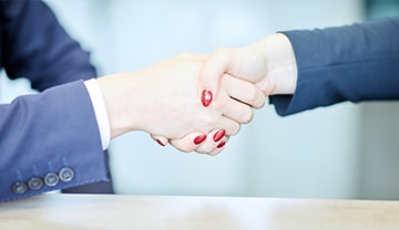 Zwei Personen in Geschäftskleidung schütteln sich an einem Tisch die Hände und symbolisieren damit eine Vereinbarung oder Partnerschaft. Der Fokus liegt auf ihren Händen, der Hintergrund ist unscharf.