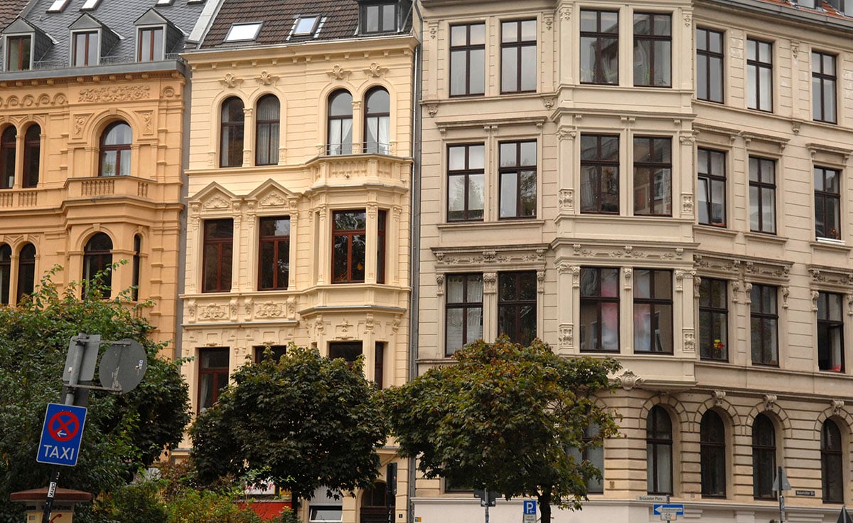Straßenansicht von eleganten, historischen europäischen Gebäuden mit verzierten Fassaden und großen Fenstern. Bäume säumen den Bürgersteig, und im Vordergrund ist ein blaues Taxiparkschild zu sehen.