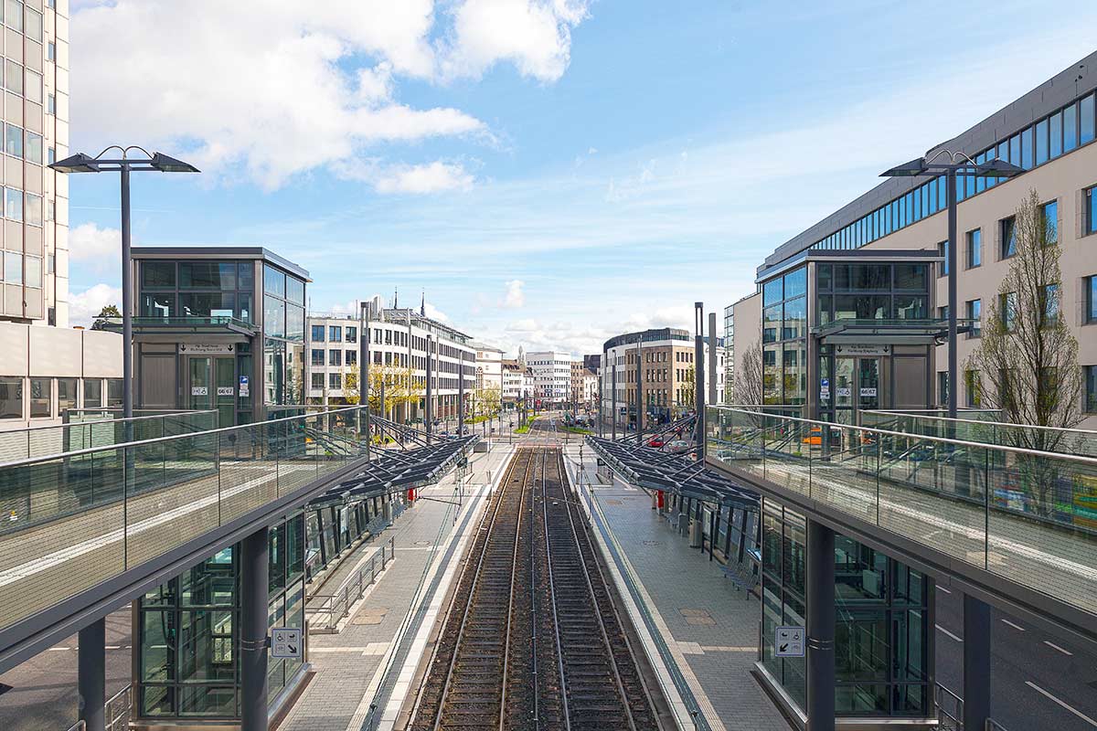 Moderner städtischer Bahnhof mit zwei Gleisen, die zwischen gläsernen Bahnsteigen verlaufen, umgeben von modernen Bürogebäuden unter einem blauen Himmel mit vereinzelten Wolken.