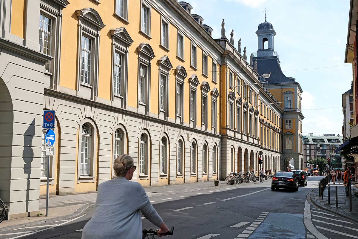 Eine Frau fährt mit dem Fahrrad auf einer Straße, an der ein großes gelbes historisches Gebäude mit Bogenfenstern steht. Entlang der Straße sind Autos und Fahrräder geparkt, und im Hintergrund erhebt sich ein Kirchturm unter einem teilweise bewölkten Himmel.