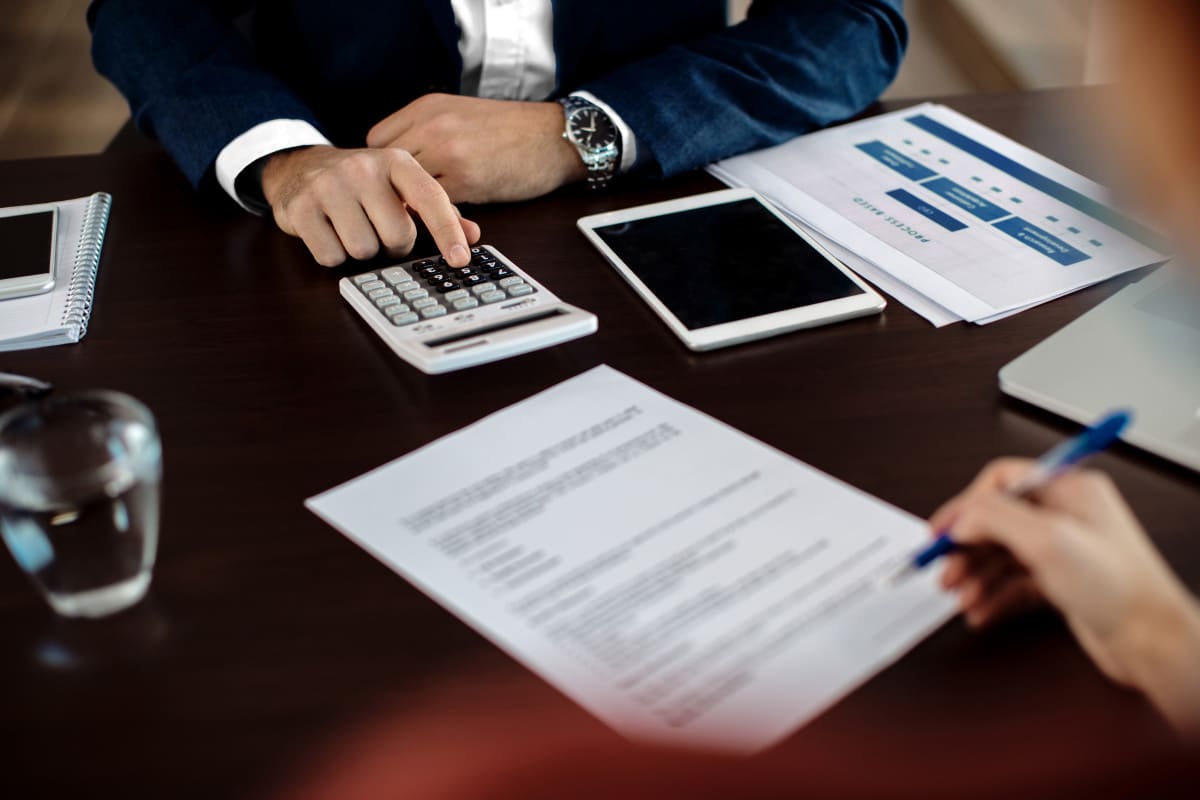 Zwei Personen arbeiten an einem Schreibtisch mit Dokumenten, einem Taschenrechner, einem Tablet und gedruckten Tabellen; eine Person zeigt auf den Taschenrechner, während die andere auf ein Papier schreibt.