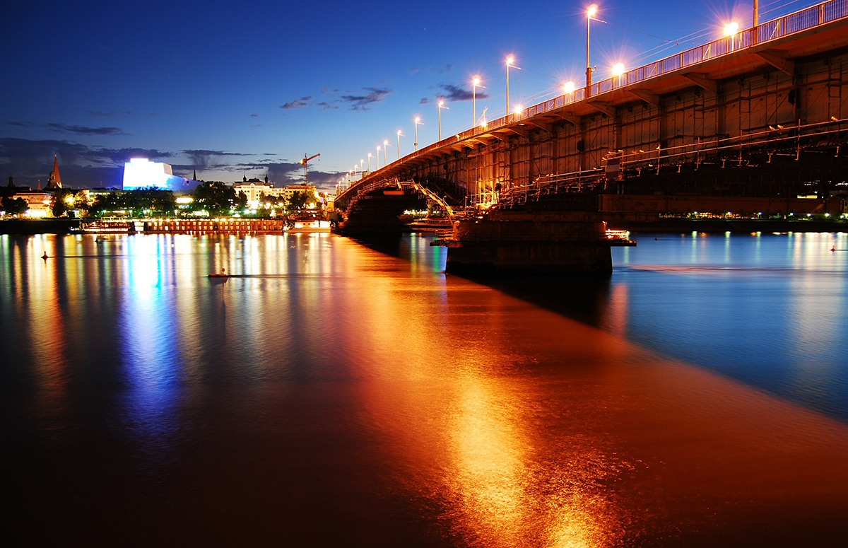 Eine von Straßenlaternen beleuchtete Brücke spannt sich in der Abenddämmerung über einen ruhigen Fluss, wobei die Lichter auf dem Wasser schimmern und im Hintergrund eine Stadtlandschaft zu sehen ist.
