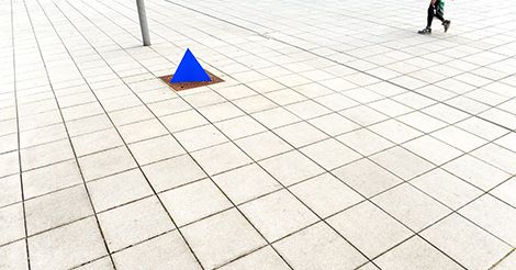 Ein blaues dreieckiges Prisma steht auf einem gefliesten, offenen Platz. In der oberen rechten Ecke geht eine Person vorbei, die größtenteils außerhalb des Bildausschnitts liegt und von der Taille abwärts sichtbar ist.