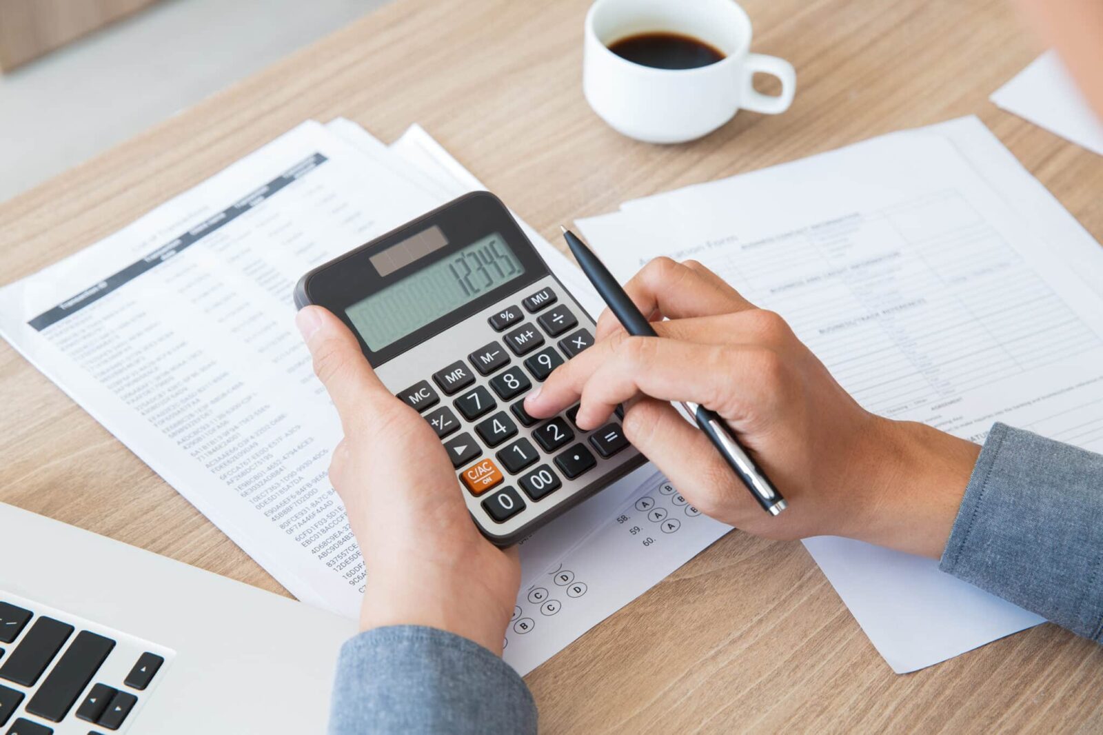 Eine Person benutzt einen Taschenrechner, während sie einen Stift in der Hand hält, mit Finanzunterlagen, einem Laptop und einer Tasse Kaffee auf einem Holztisch.