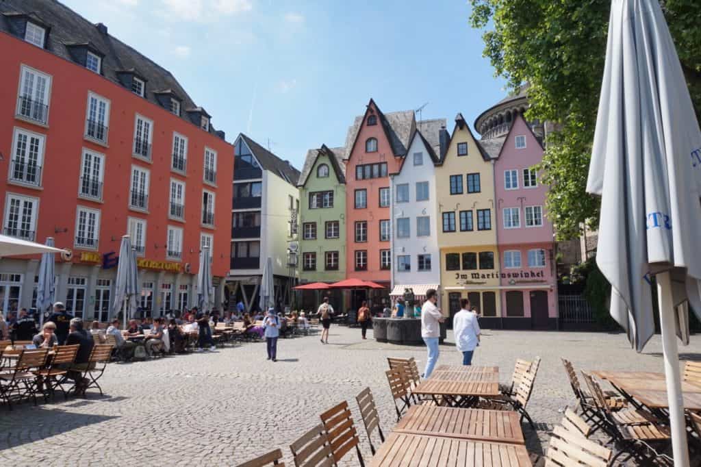 Ein sonniger Platz mit bunten, schmalen Gebäuden, Caféterrassen und einigen Passanten spiegelt den Charme wider, den Makler in Köln vermitteln, wenn sie Immobilien vor dem Verkauf professionell in Szene setzen. Auf der rechten Seite säumen Bäume den belebten Platz.