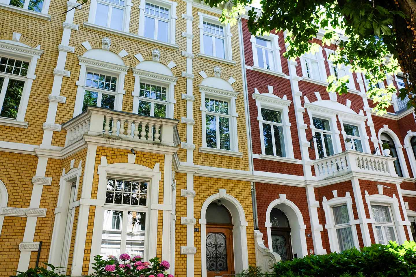 Ein Blick auf bunte Backstein-Stadthäuser mit gewölbten Türen, weißen Zierleisten, großen Fenstern und Balkonen, die teilweise von grünen Bäumen beschattet und von blühenden Sträuchern im Vordergrund eingerahmt werden.