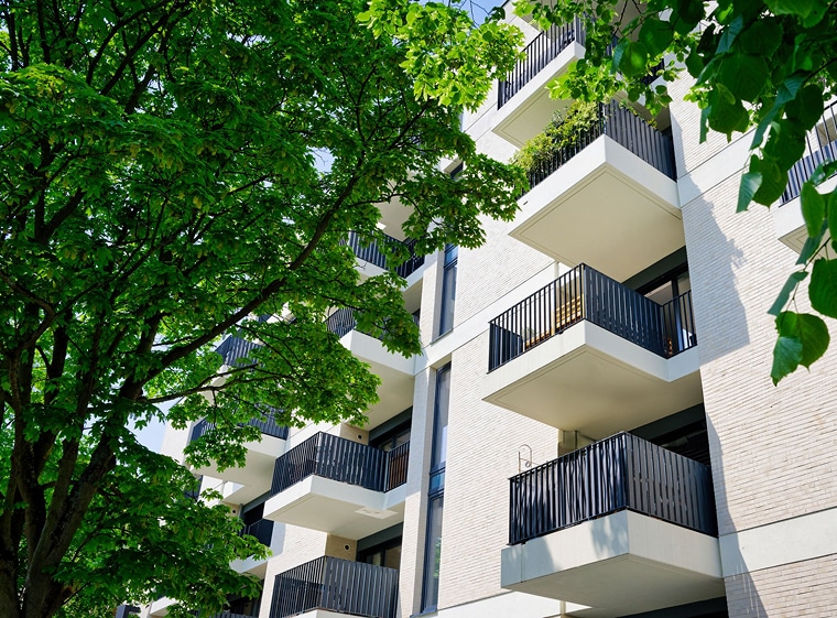 Modernes Mehrfamilienhaus mit mehreren Balkonen und schwarzen Geländern, die an einem sonnigen Tag von grünen, belaubten Bäumen im Vordergrund teilweise beschattet werden.