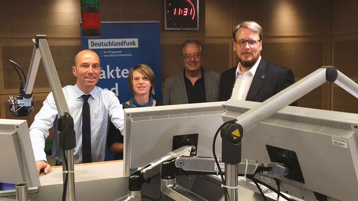 Vier Personen stehen hinter Pulten mit Monitoren und Mikrofonen in einem Radiostudio. Im Hintergrund sind ein Deutschlandfunk-Schild und eine Uhr, die 11:31 Uhr anzeigt, zu sehen. Die Gruppe lächelt und ist in Business-Kleidung gekleidet.