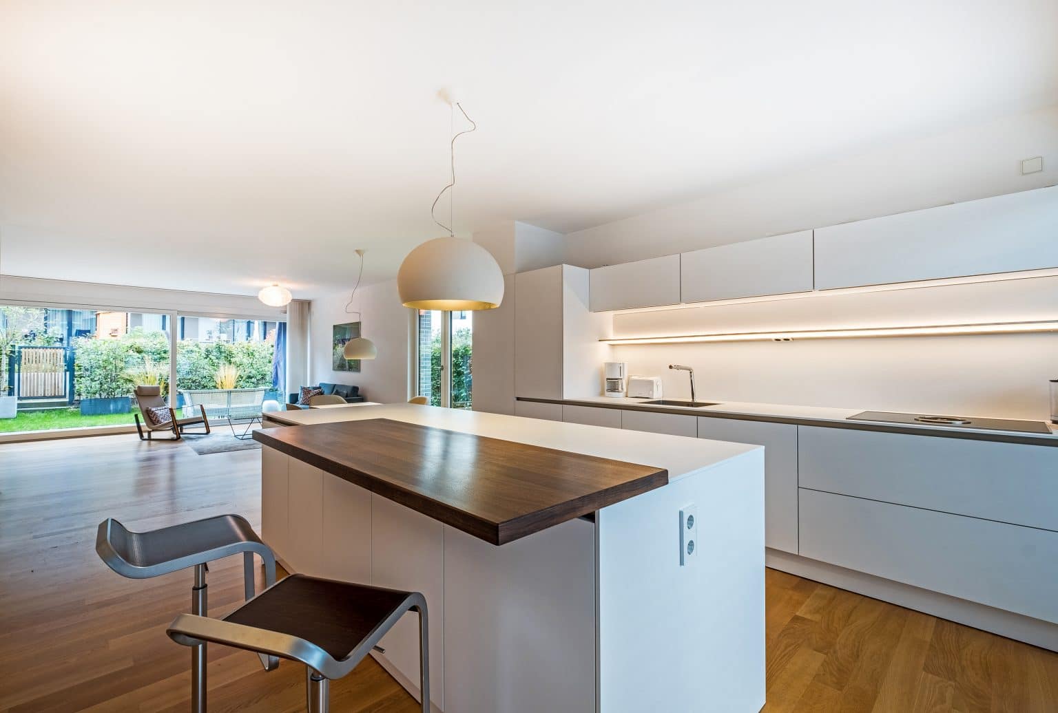 Moderne, offene Küche mit weißen Schränken, Holzinsel, Barhocker, Hängelampen und großen Fenstern, die zum hellen Wohnbereich mit Gartenblick führen.