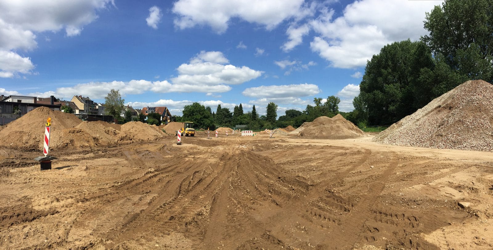 Eine große, schlammige Baustelle mit Haufen von Erde, Schotter, Warnschildern und Reifenspuren. Im Hintergrund sind Bäume und Häuser unter einem teilweise bewölkten Himmel zu sehen.