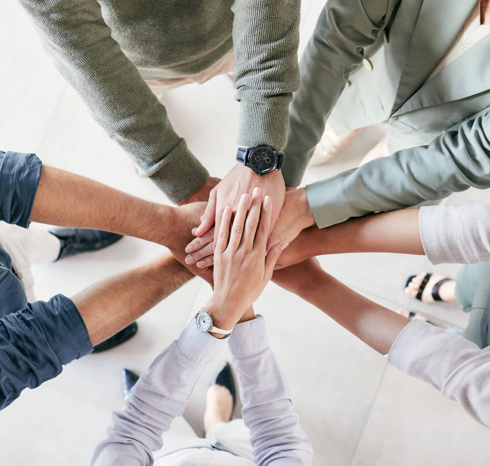 Eine Gruppe von Menschen, die in einem Kreis stehen, legen ihre Hände in der Mitte zusammen und symbolisieren so Teamwork und Einheit. Das Foto ist von oben aufgenommen und zeigt nur ihre Arme und Hände.