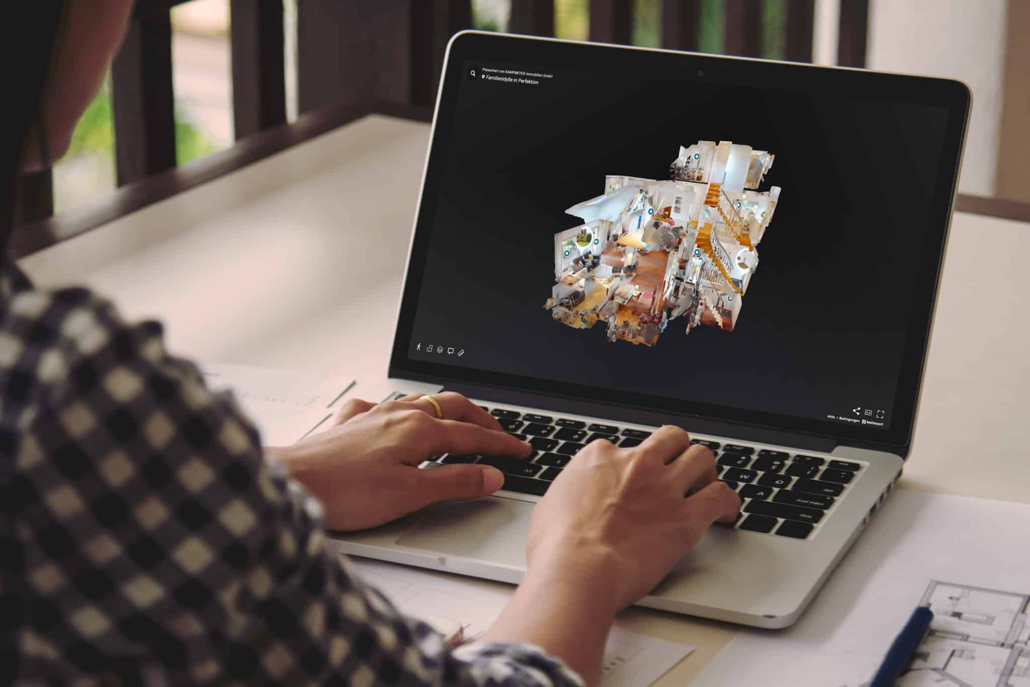 Eine Person, die einen Laptop benutzt, betrachtet ein digitales 3D-Modell eines Gebäudeinneren auf dem Bildschirm, während in der Nähe Architekturpläne und Unterlagen auf dem Schreibtisch liegen.