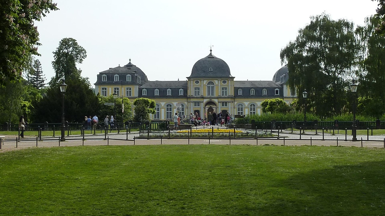 Unter strahlend blauem Himmel versammeln sich Menschen vor einem großen, gelben, historischen Gebäude mit Kuppeldächern, das von Gärten und Rasenflächen umgeben ist – und diskutieren vielleicht Fragen wie: Was ist besser: Eine Immobilie in Bonn zu verkaufen oder zu vermieten?.