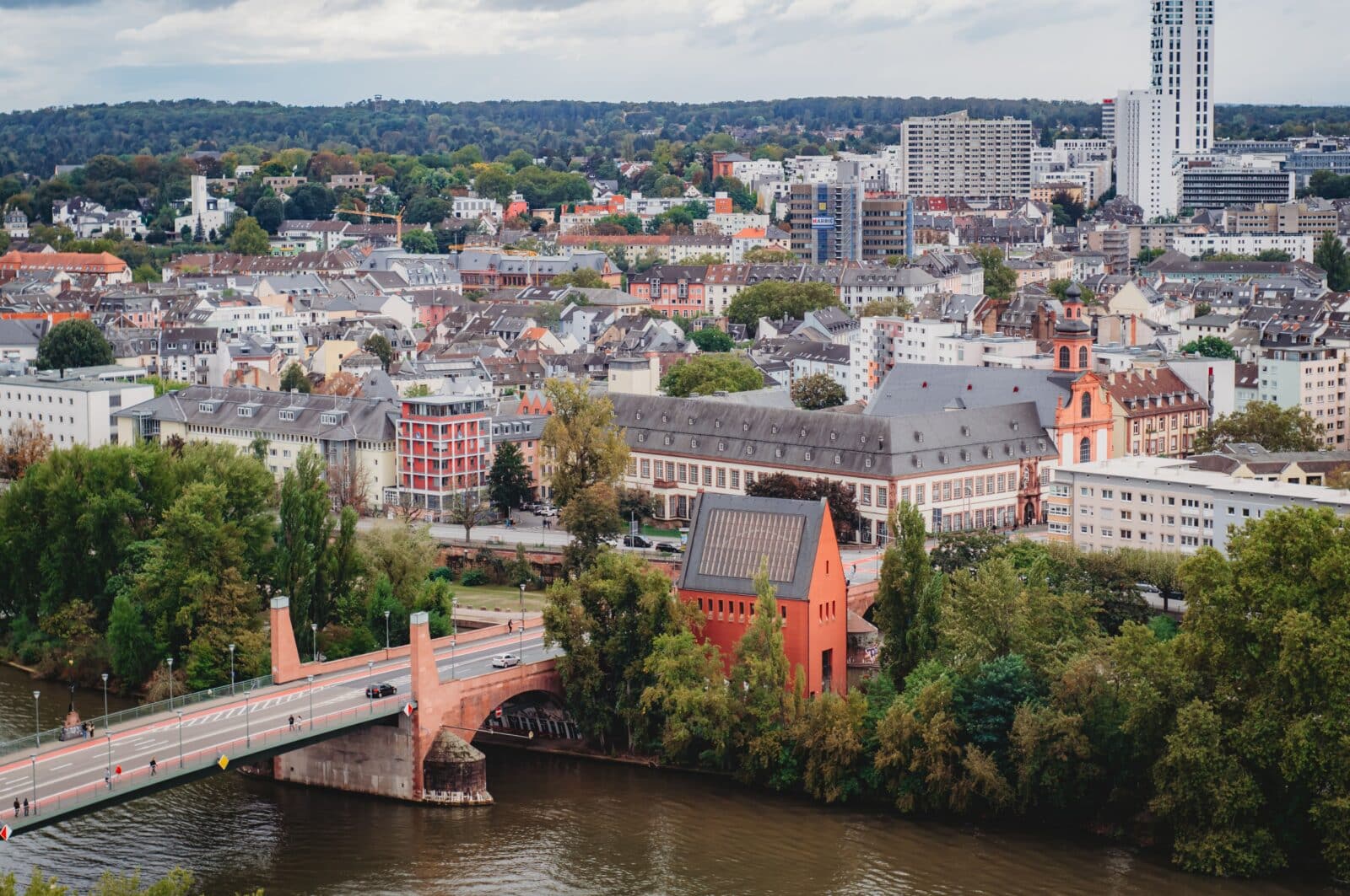 Ein Stadtbild zeigt eine Brücke, die einen Fluss überspannt, Bäume, die das Ufer säumen, sowie eine Mischung aus modernen und historischen Gebäuden mit einem hohen weißen Wolkenkratzer im Hintergrund – eine Atmosphäre, die die dynamische Immobilienlandschaft widerspiegelt, in der diejenigen, die sich fragen: „Welche Immobilienmakler in Bonn sind auf Erbimmobilien spezialisiert?“, in der vielfältigen Architektur der Stadt Inspiration finden könnten.