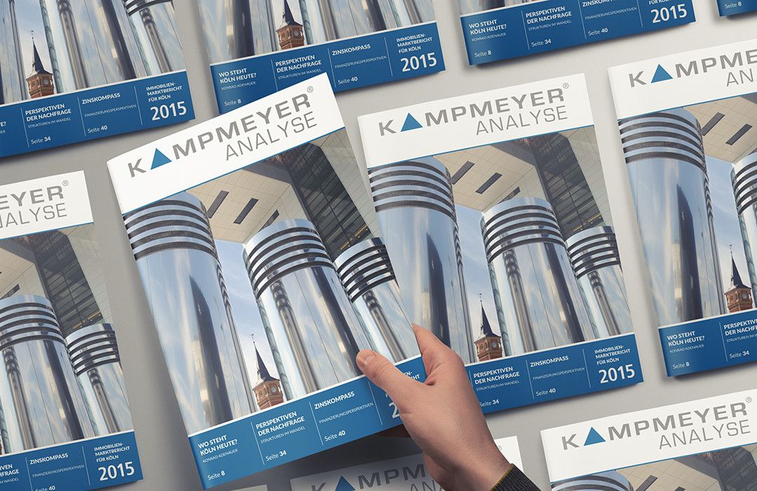 Eine Hand nimmt eine Zeitschrift KAMPMEYER ANALYSE 2015 in die Hand, die ein Titelbild mit modernen spiegelnden Gebäuden und einem Kirchturm zeigt, umgeben von mehreren Exemplaren derselben Zeitschrift.